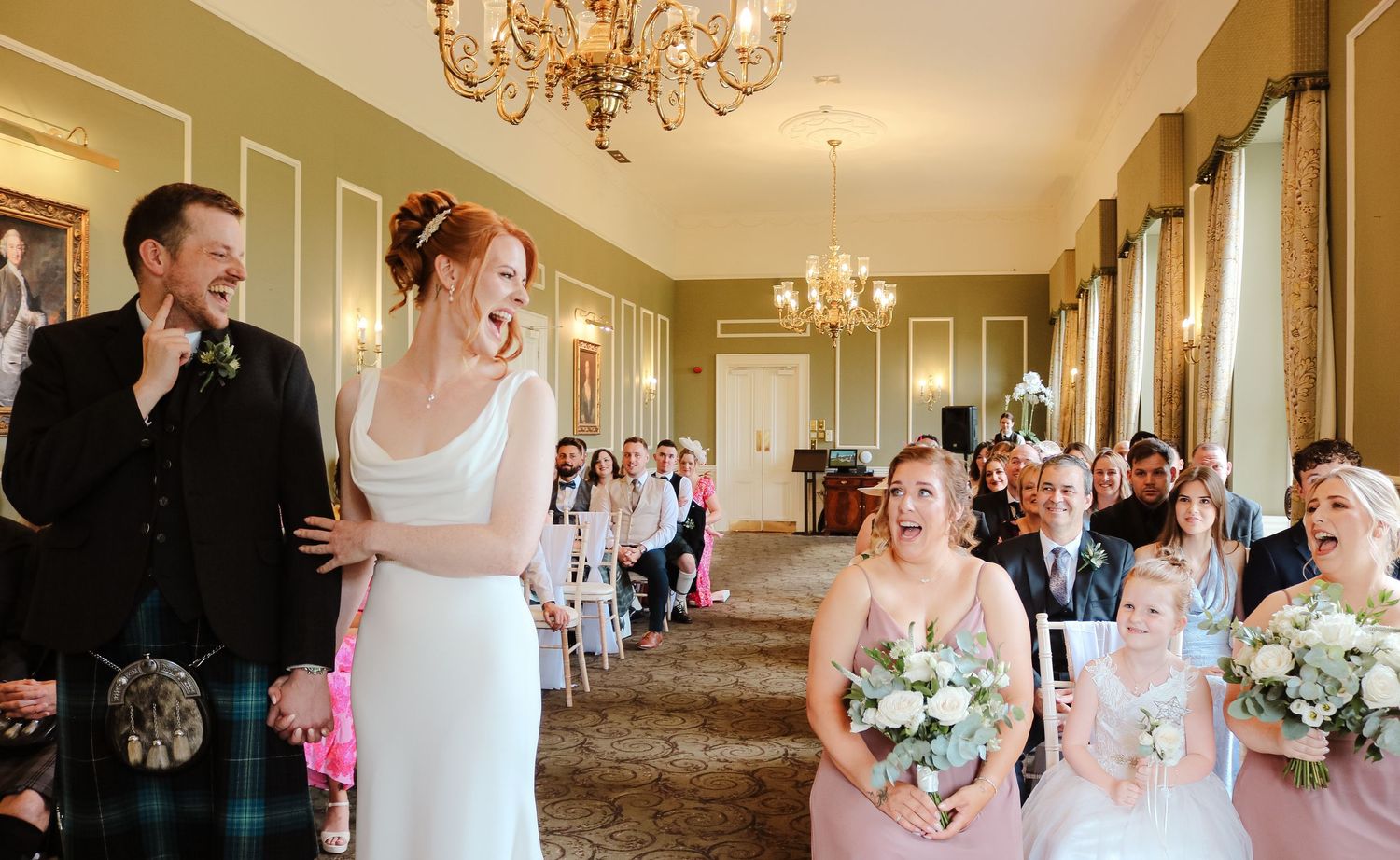 An elegant wedding ceremony taking place in a chandelier-lit corridor with classic green walls and tall windows.