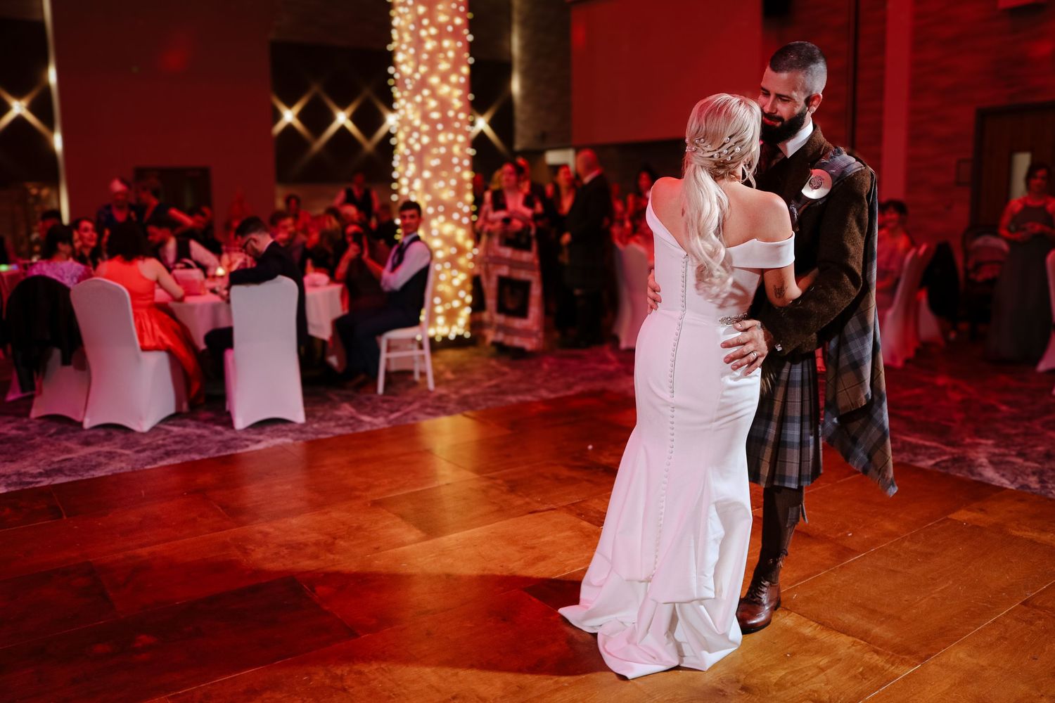 A couple shares their first dance at a wedding reception in a red-lit ballroom with white chairs and twinkle lights.