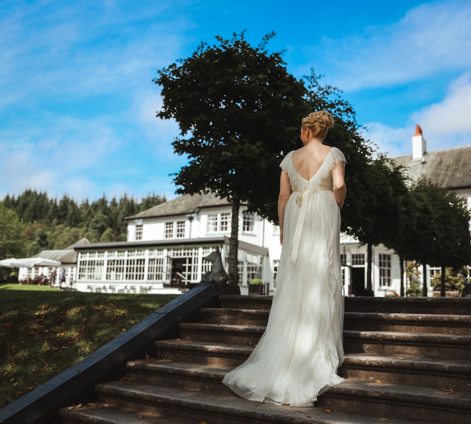 A person in an elegant white wedding dress stands on stone steps in front of a large white country house on a sunny day.