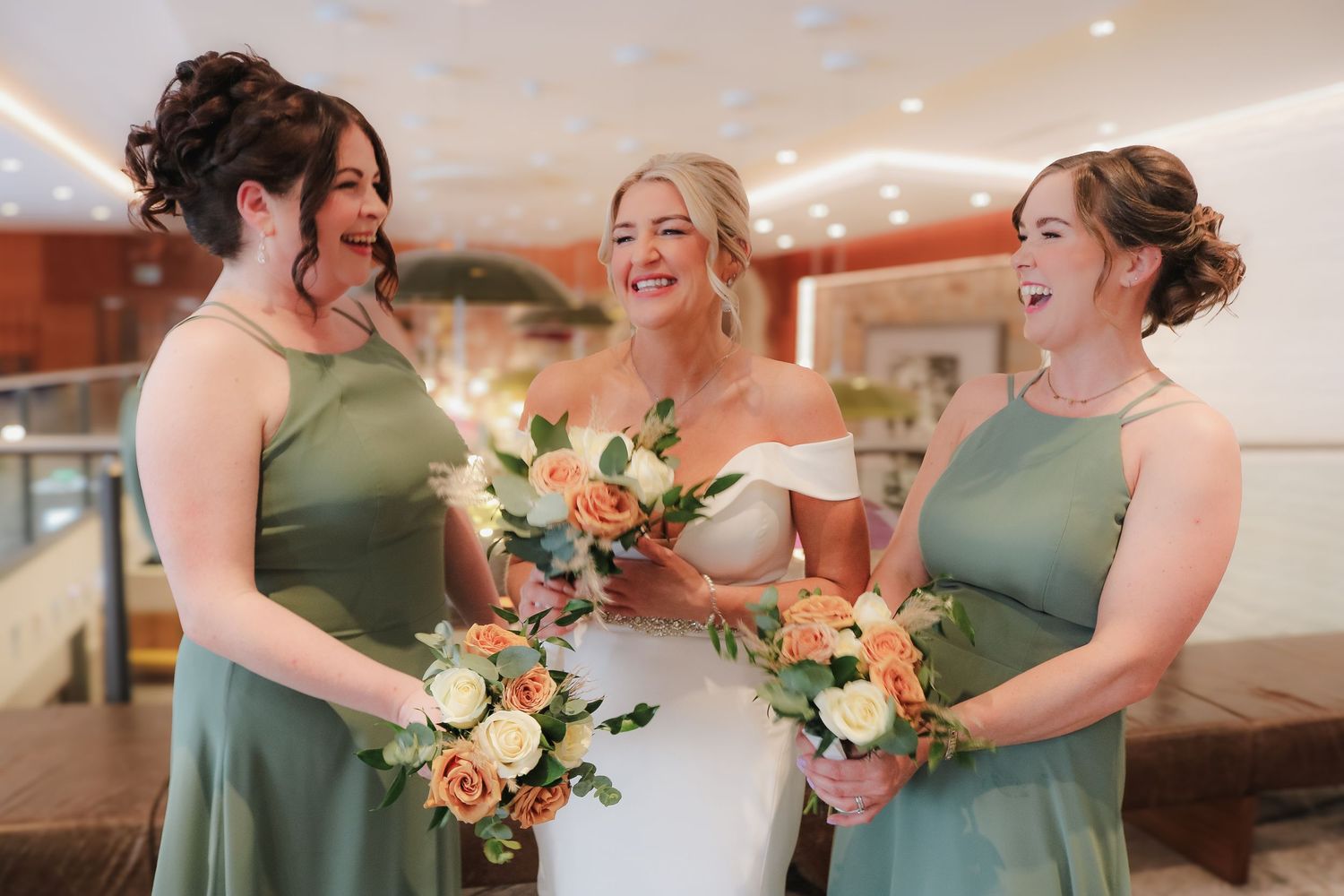 A group of bridesmaids in green dresses share joyful moments and hold peach-colored floral bouquets at a wedding.