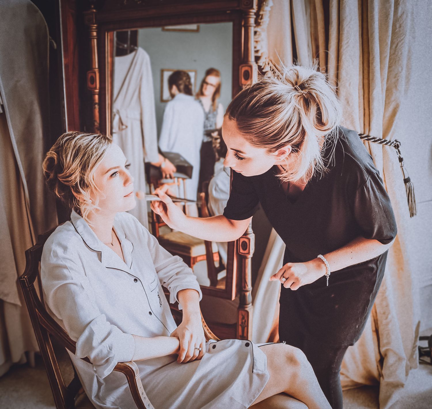 Makeup artist applying cosmetics to a client seated in a dressing room with hanging clothes in the background.