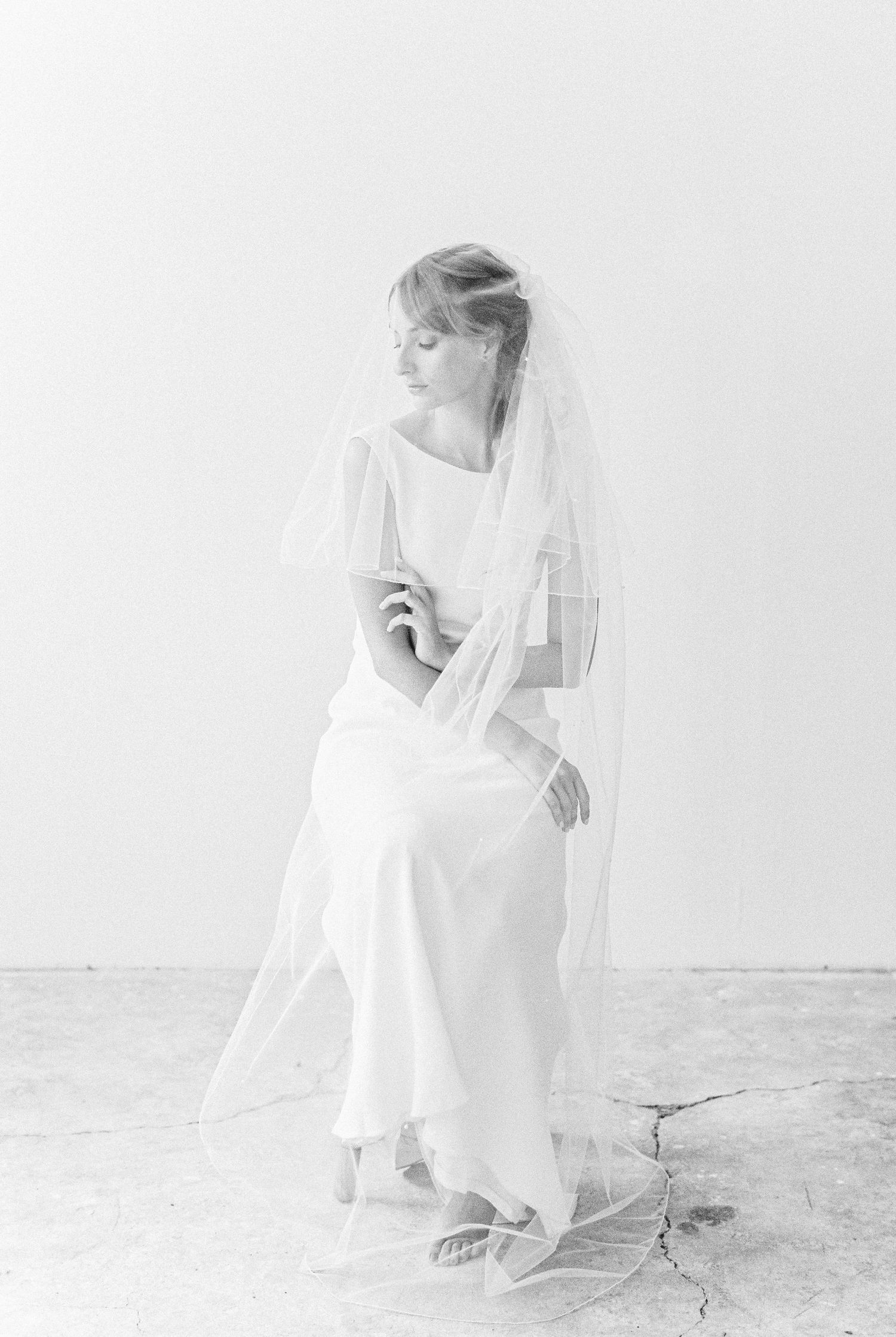 Ethereal black and white bridal portrait of a figure in a flowing white dress and veil against a minimalist background.