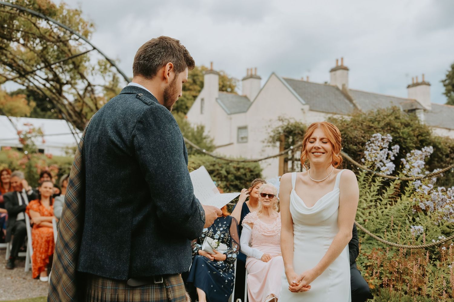 People gather at an outdoor wedding ceremony on a sunny day, with white buildings and greenery in the background.