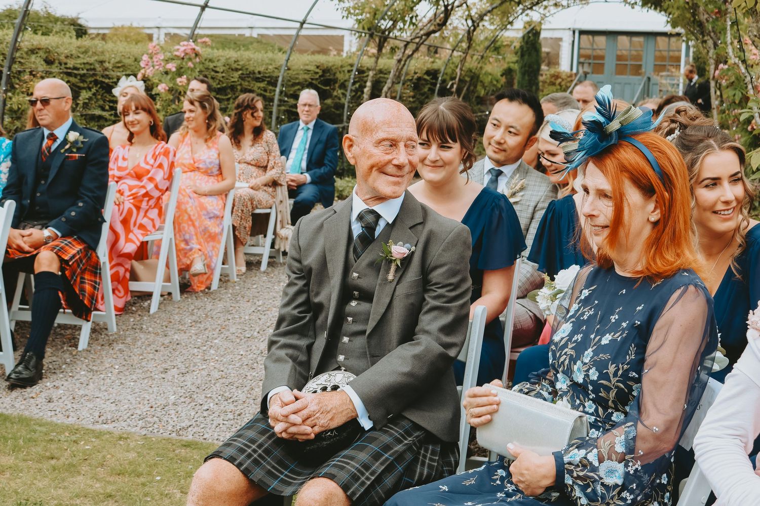 Wedding guests seated outdoors for ceremony with person in traditional Scottish kilt in foreground.