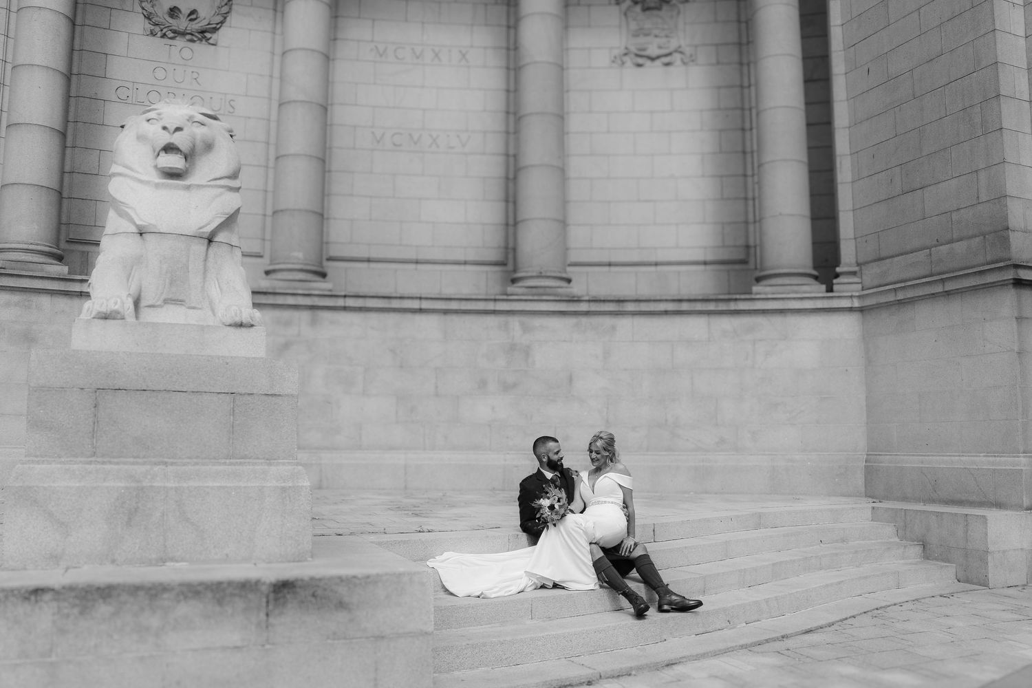 People sit on the steps of a grand building with ornate stone architecture and decorative sculptural elements.