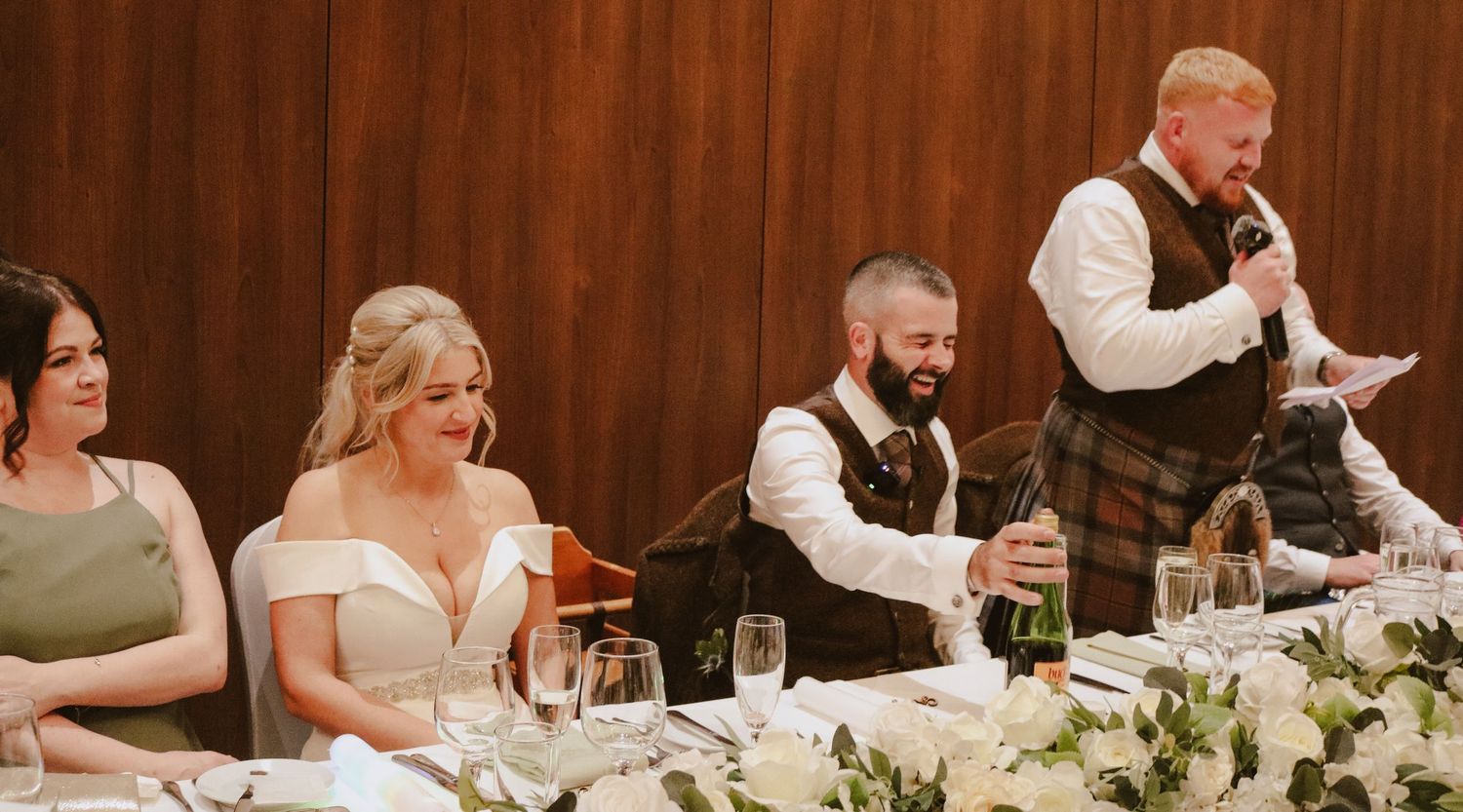 Wedding reception guests seated at a long rustic table with white floral centerpieces and elegant place settings.