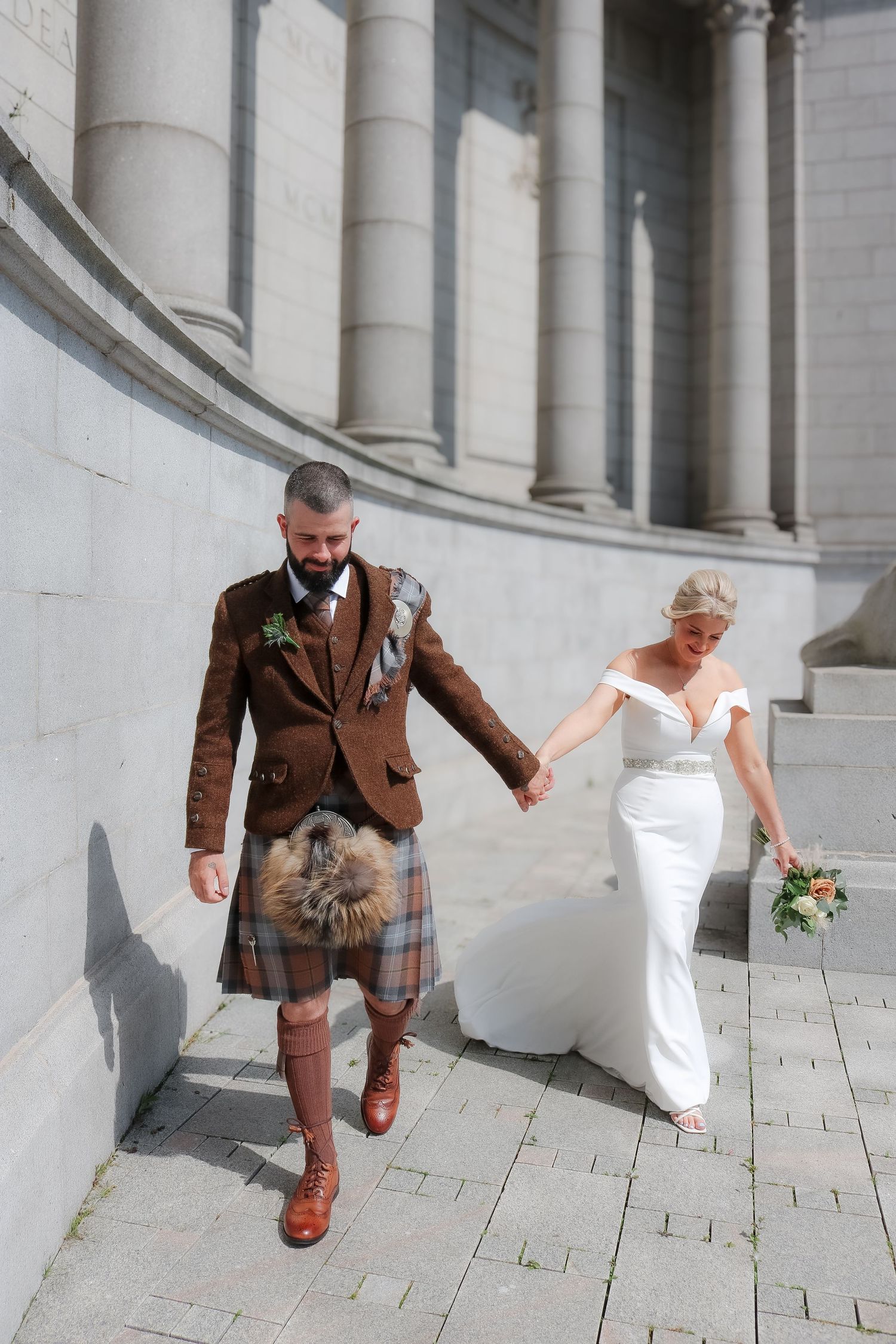 A couple in wedding attire walking against a white columned building, with the groom wearing a traditional Scottish kilt.