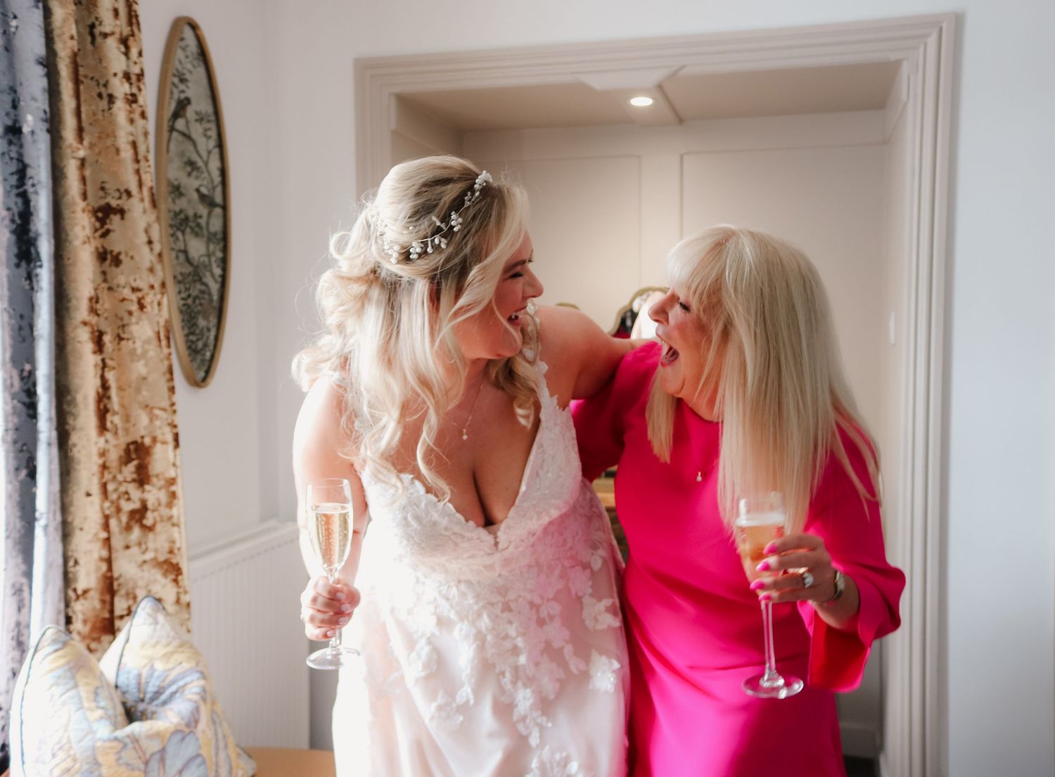 Two women sharing a joyful moment and laughter while holding champagne glasses in an elegant white and pink setting.