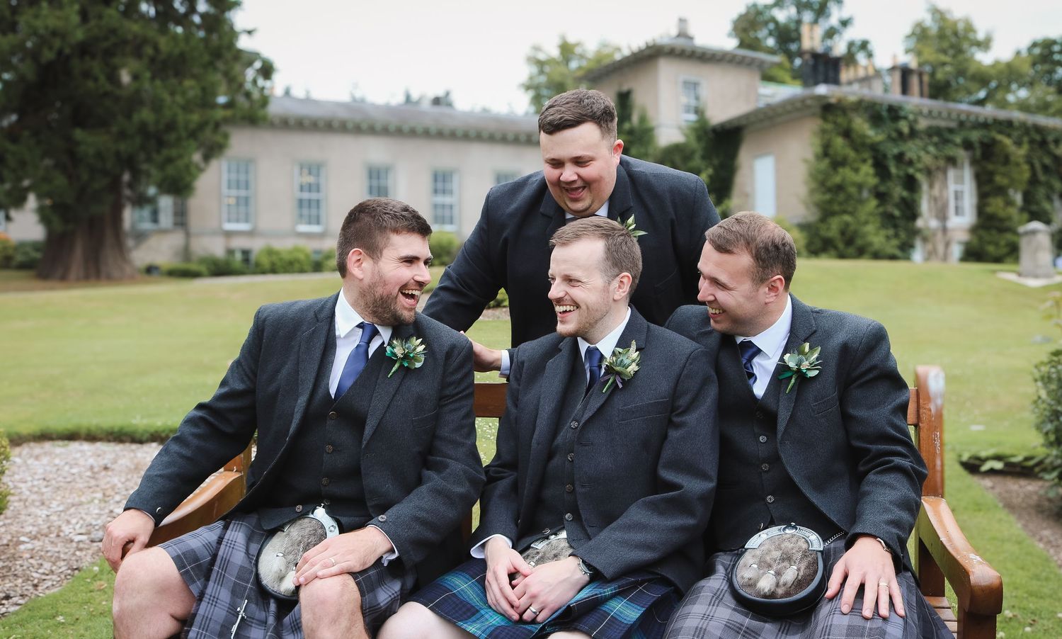 A joyful group in formal black suits laughs and jokes together while sitting outdoors on a grassy lawn.