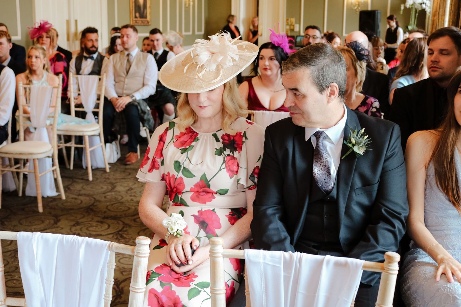Wedding guests seated in white chairs at an elegant indoor ceremony venue with floral decor details.