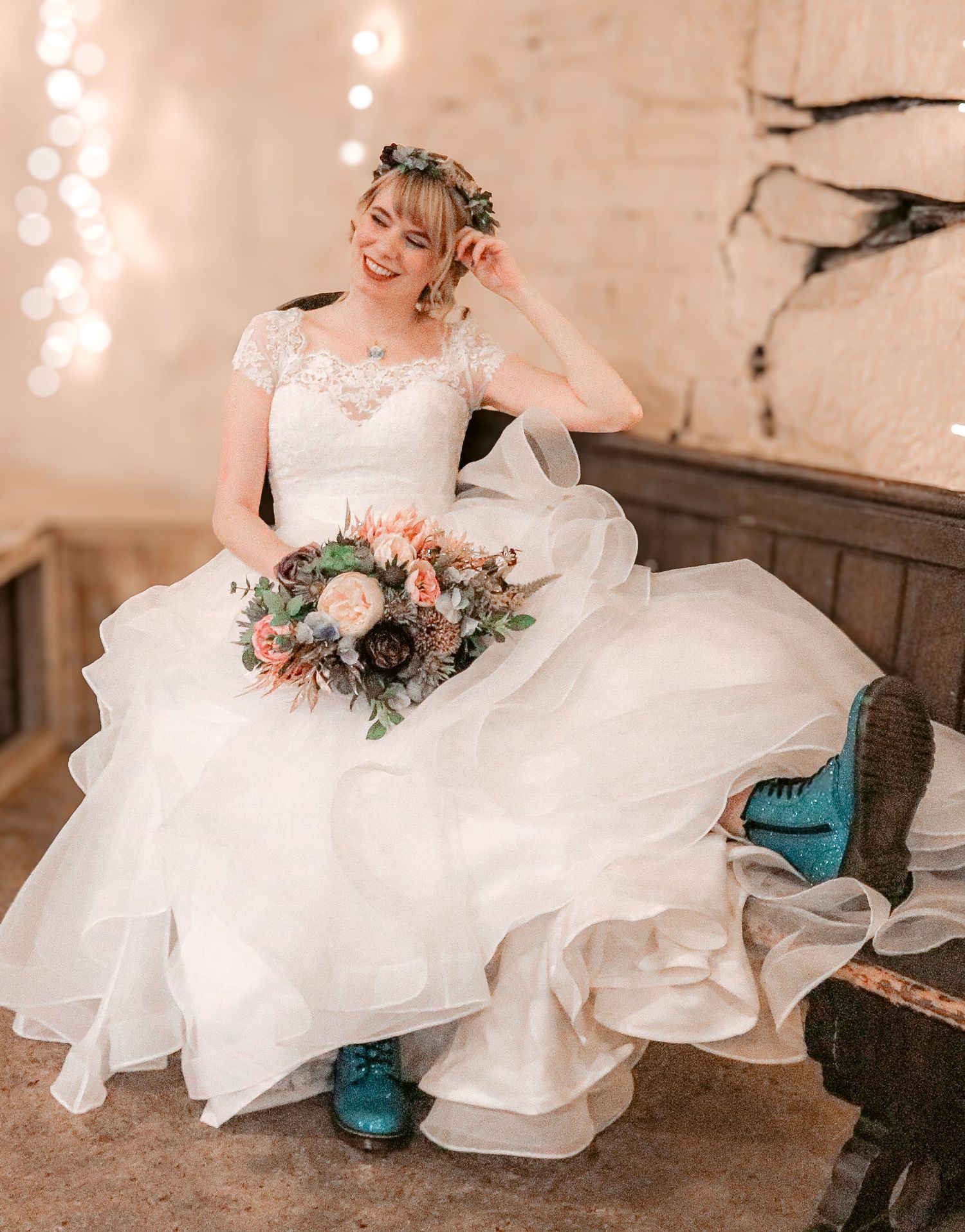 A bride in an elegant off-shoulder white gown sits elegantly with a pastel floral bouquet against a rustic backdrop with lights.