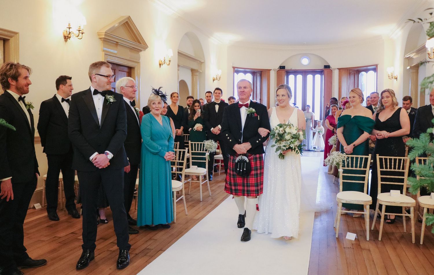 Wedding ceremony at an elegant venue with a groom in traditional Scottish kilt walking down the aisle.