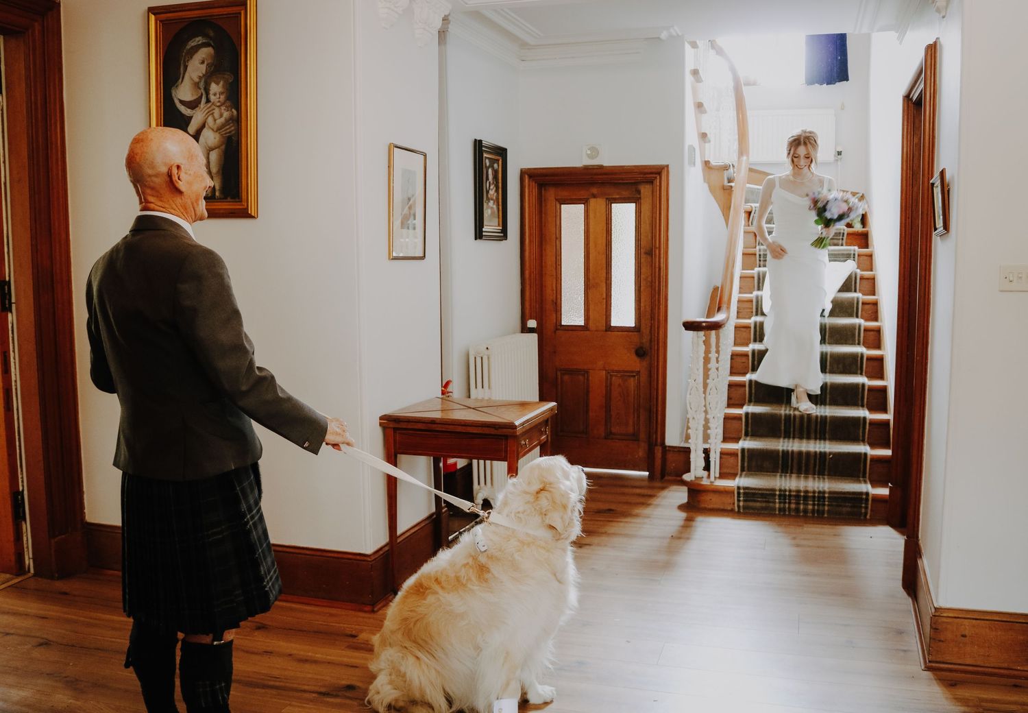 Someone stands with their Golden Retriever dog in an entryway while another figure carries flowers up wooden stairs.