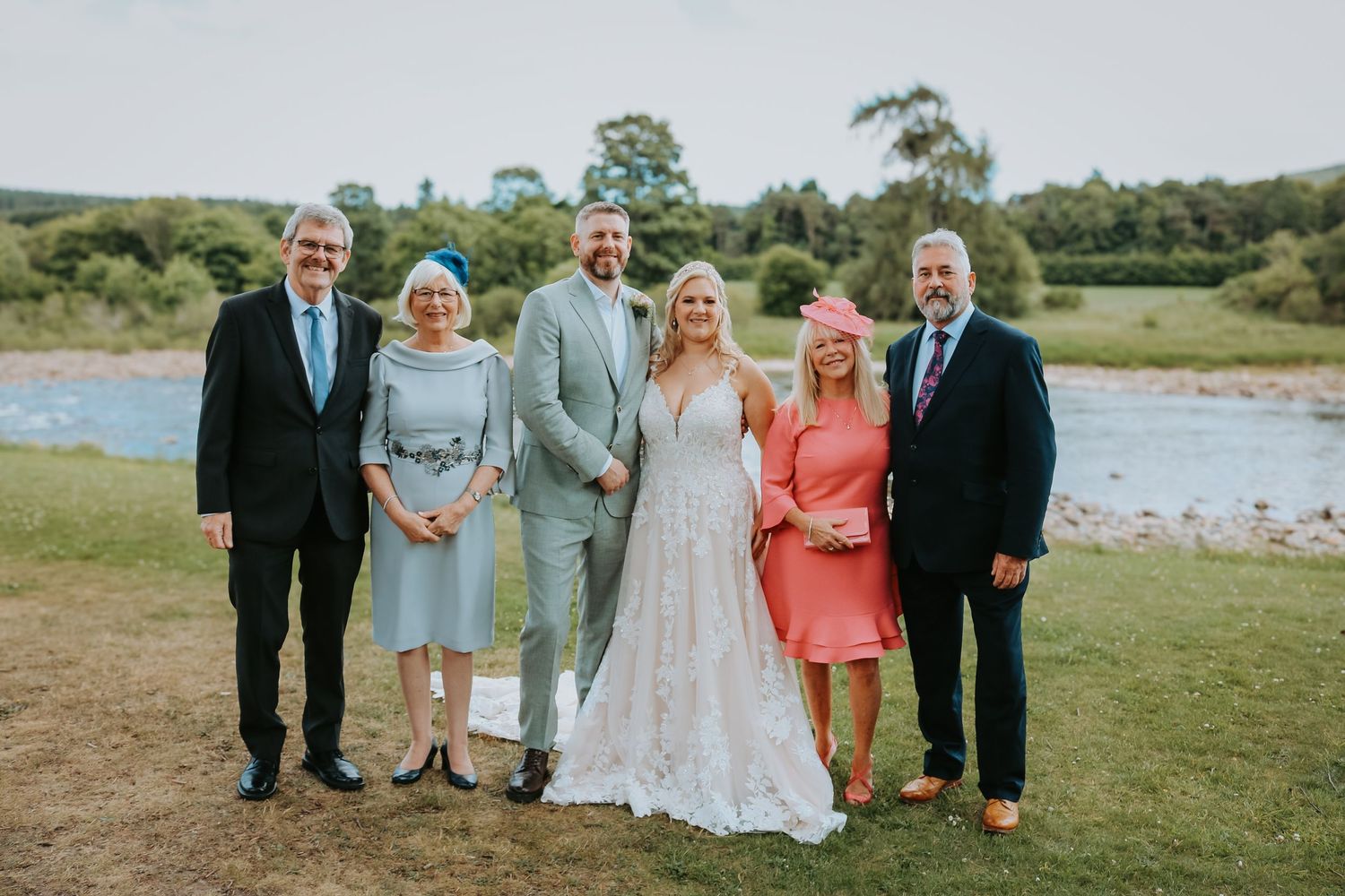 A group of formally dressed guests standing together outdoors on a green lawn near a waterfront during a wedding celebration.