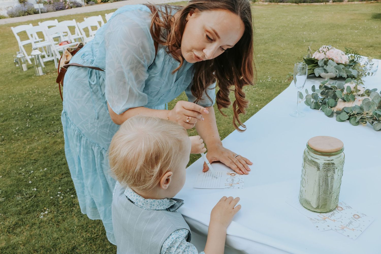 A child and caregiver work together at an outdoor table at an event with white folding chairs in the background.