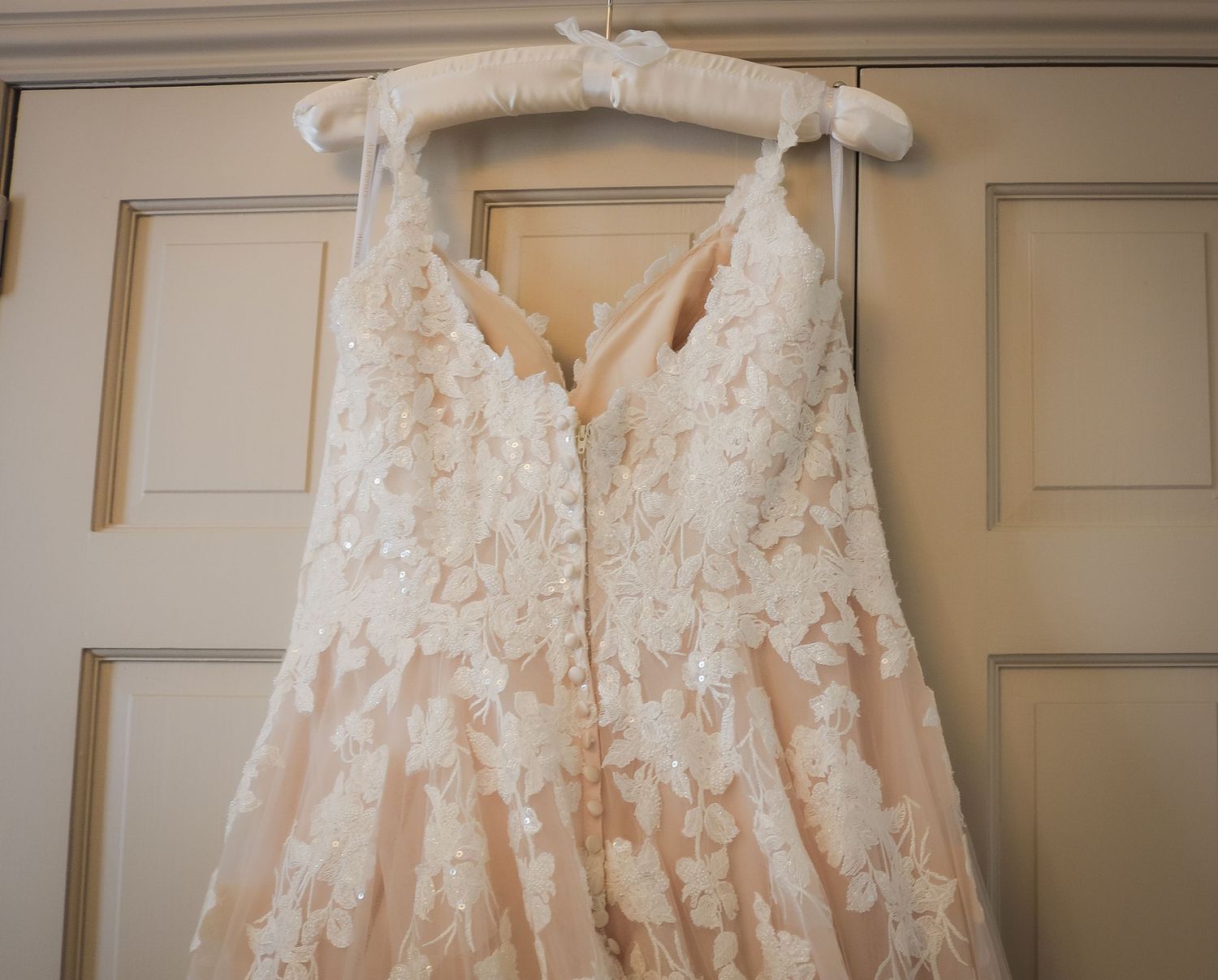 A delicate ivory lace wedding dress with floral appliques hangs on a padded hanger against a cream-colored door.