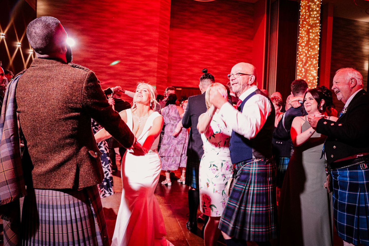 Wedding guests in traditional Scottish kilts dance together at a reception with red mood lighting and decorative backdrop.