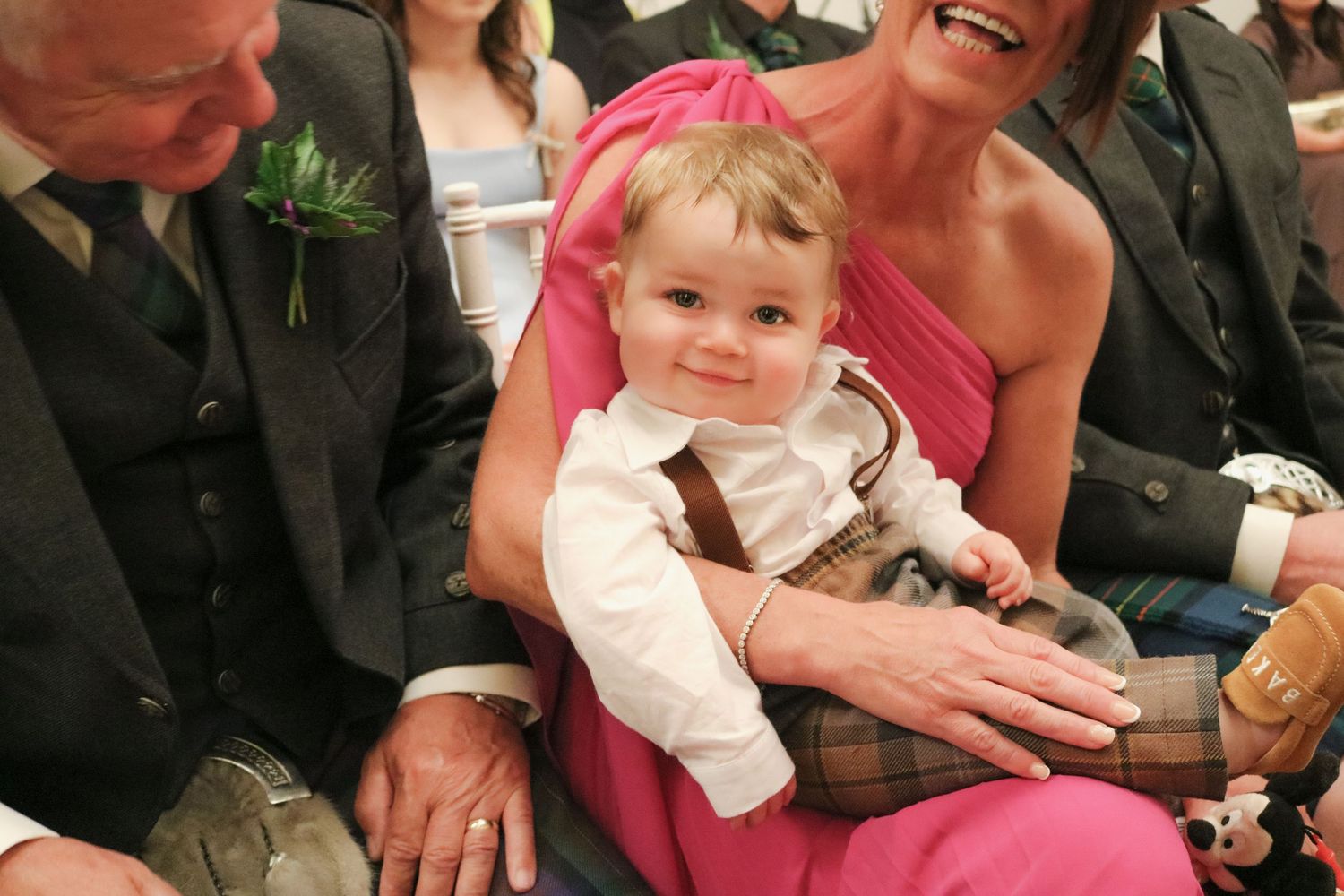 A smiling toddler in a white dress shirt sits with adults at a formal event.