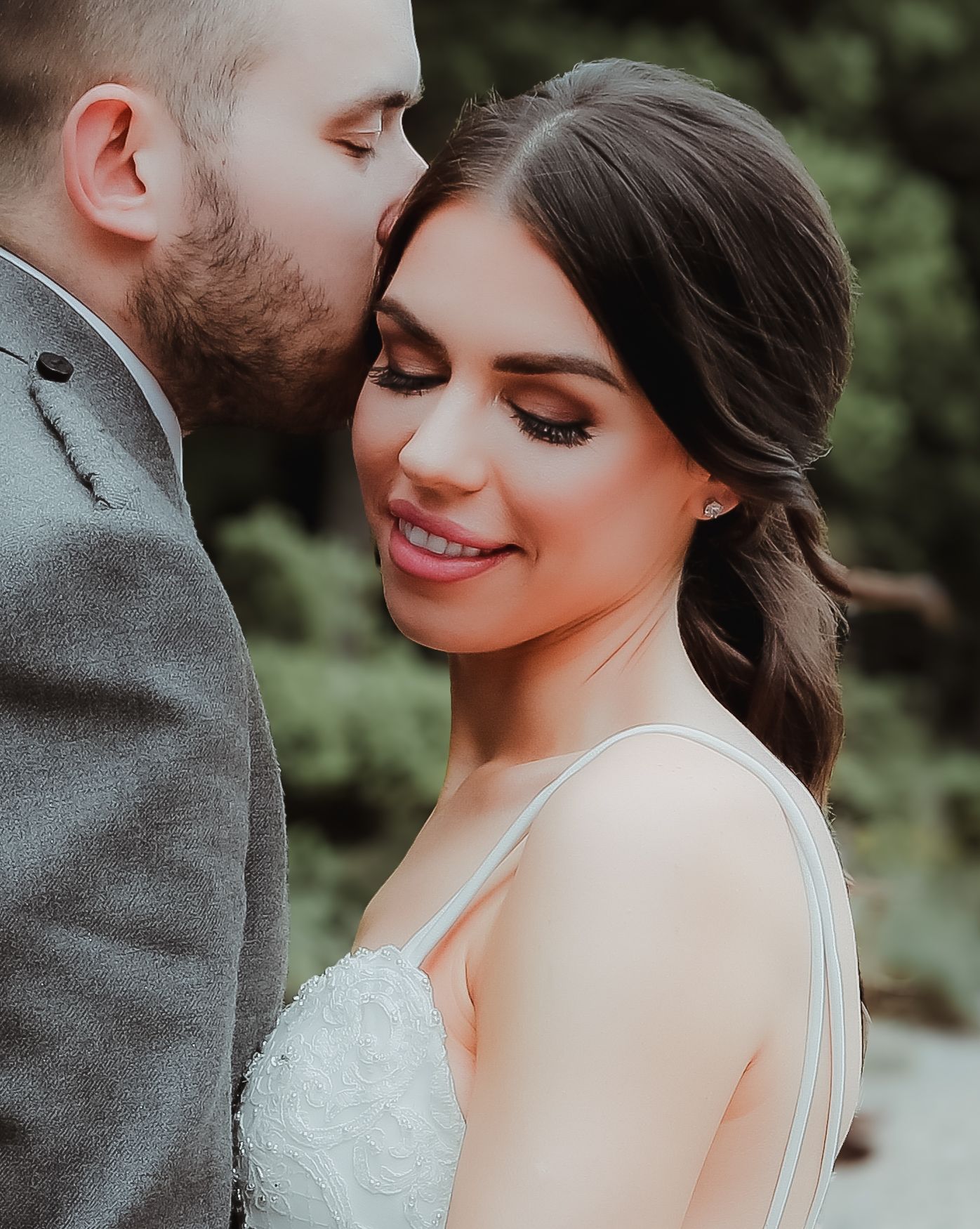 Romantic wedding photo showing close-up of bride in white lace dress smiling as groom whispers in her ear in garden setting.