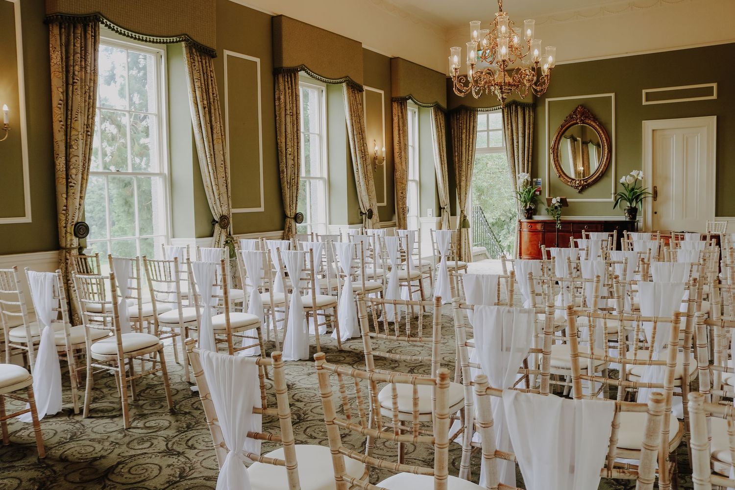 Elegant wedding venue setup with white chiavari chairs and flowing white fabric in a grand room with chandeliers.
