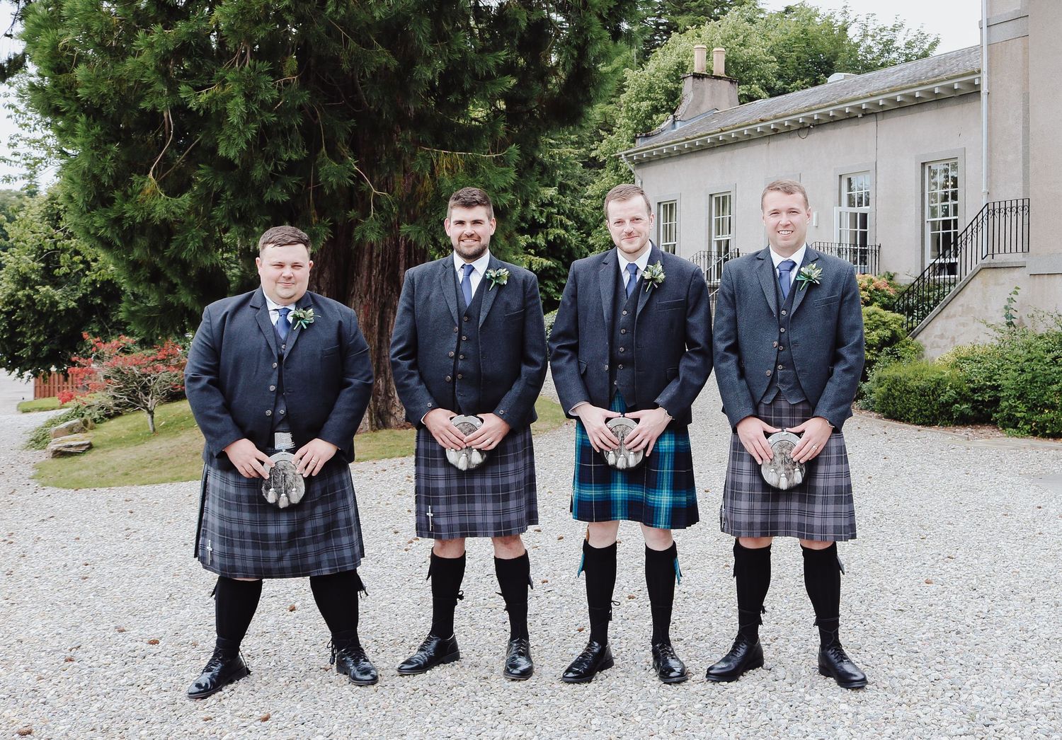 Four people wearing traditional Scottish kilts and formal attire stand together on a gravel path outside a stone building.