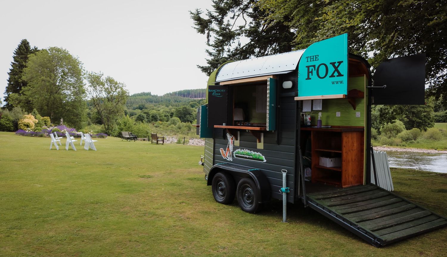 Small turquoise mobile food trailer parked on grassy lawn with Adirondack chairs visible in background.