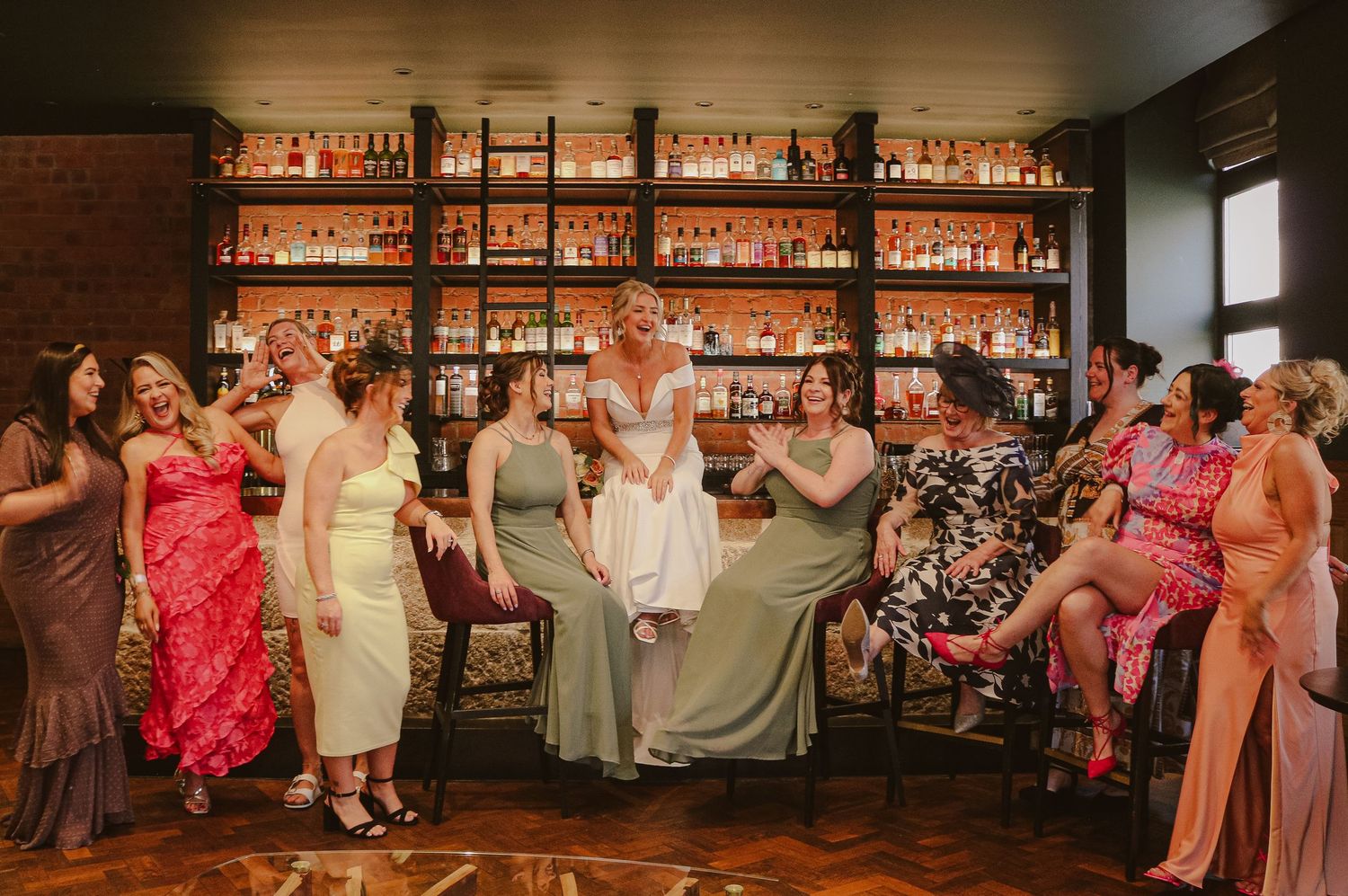 A festive group dances together at a bar with illuminated shelves of bottles glowing warmly in the background.