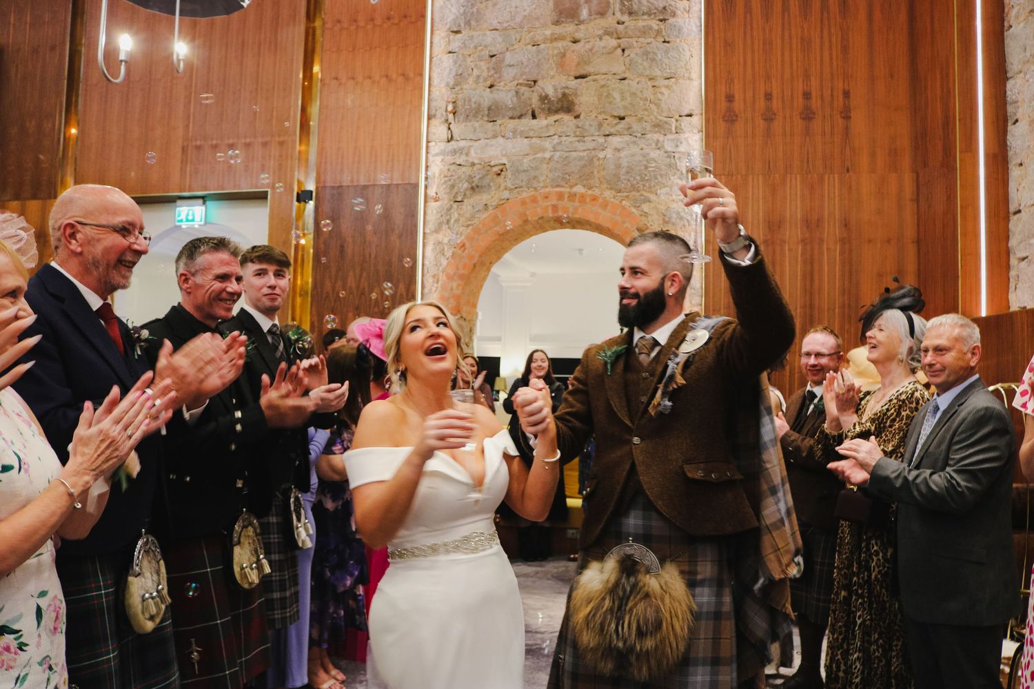 Wedding guests clap and celebrate during reception in wood-paneled venue with joyful atmosphere.