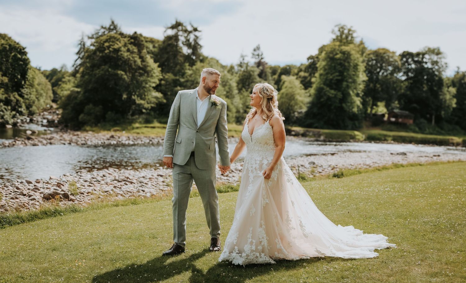 Couple in wedding attire standing together on lush green lawn beside a rocky riverbank.