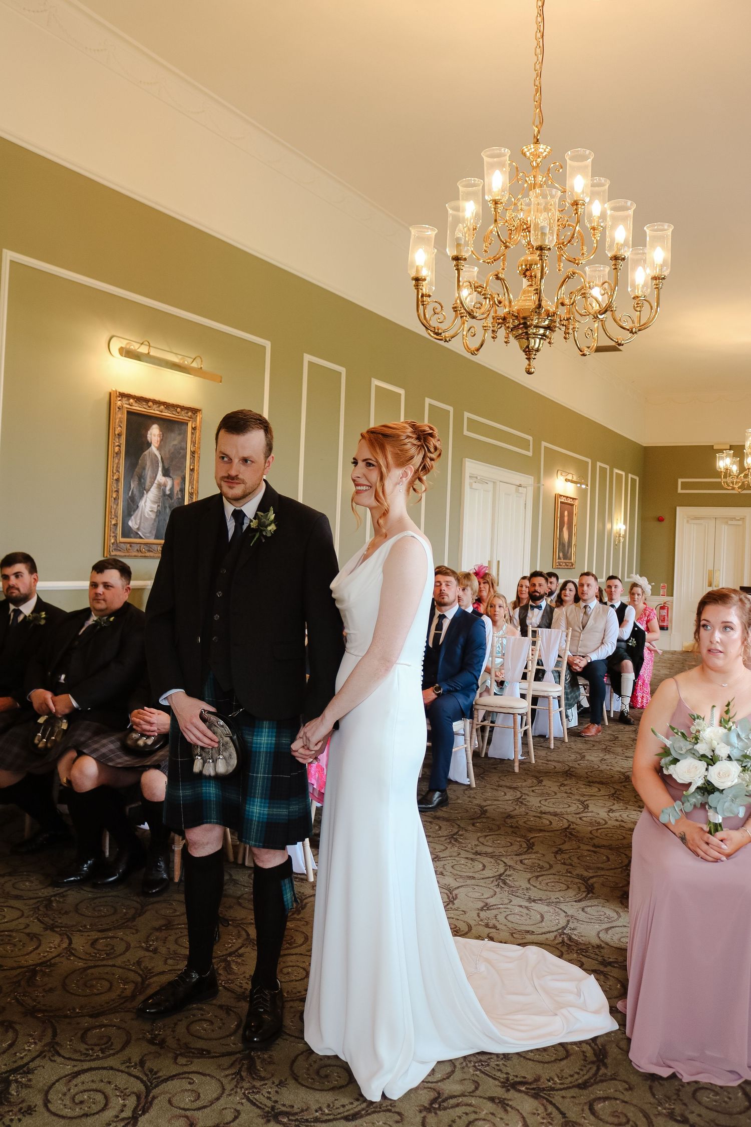 A wedding ceremony takes place in an elegant room with a crystal chandelier and green walls as attendants look on.