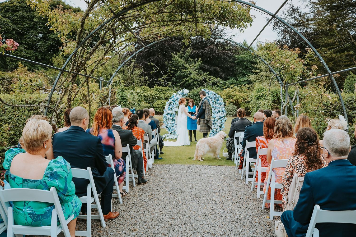 Outdoor wedding ceremony under a metal arch with guests seated on white chairs along a garden path.
