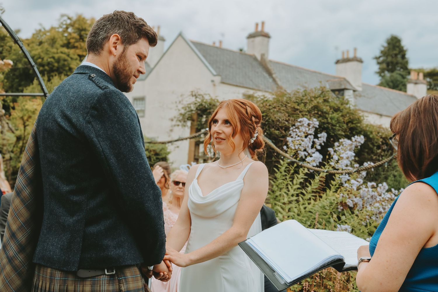 A couple exchanges vows during an intimate outdoor garden wedding ceremony with white houses in the background.