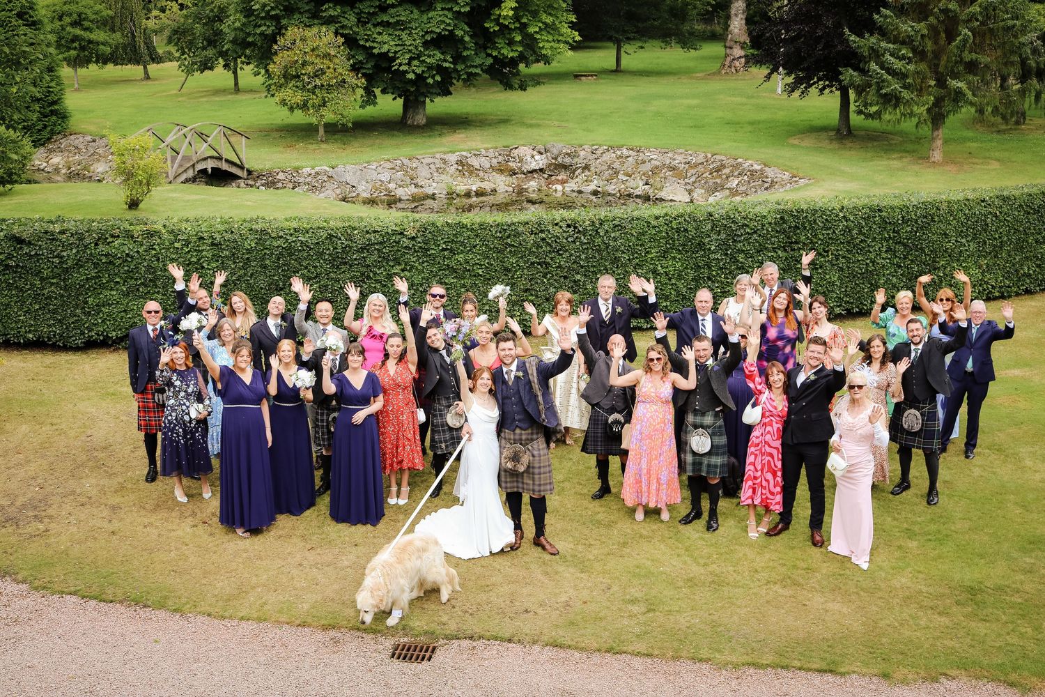Large celebration group poses for aerial photograph on green lawn with gardens and landscaping in background.