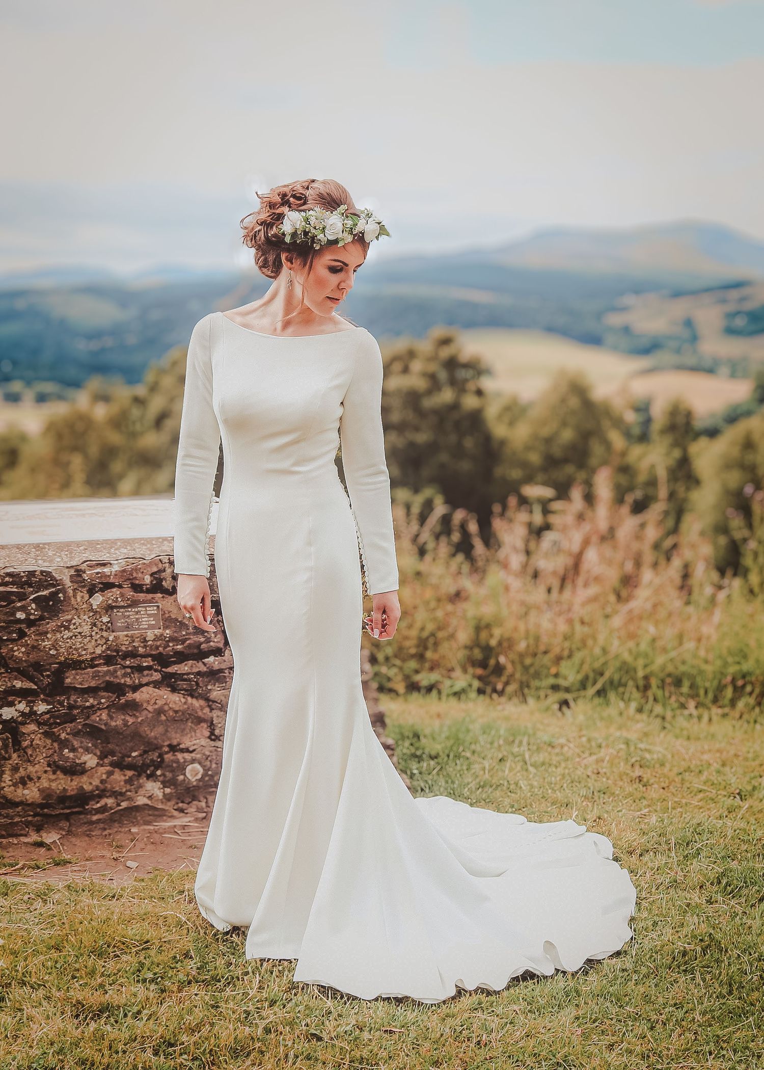 Elegant long-sleeved white wedding dress with a flowing train photographed against a scenic mountain landscape at sunset.