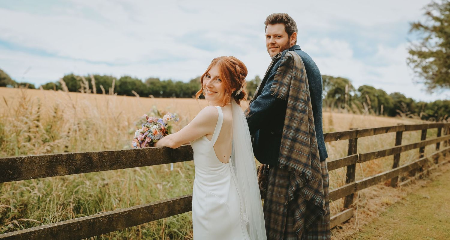 Wedding couple stands together along a wooden fence overlooking a rustic country field on a cloudy day.