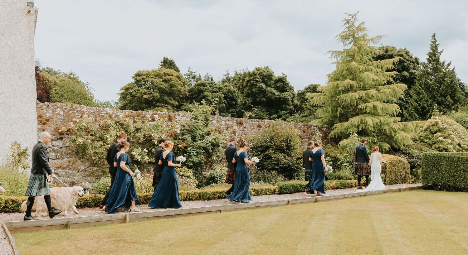 People in coordinated navy dresses joyfully jump in unison on a green lawn near a garden wall.