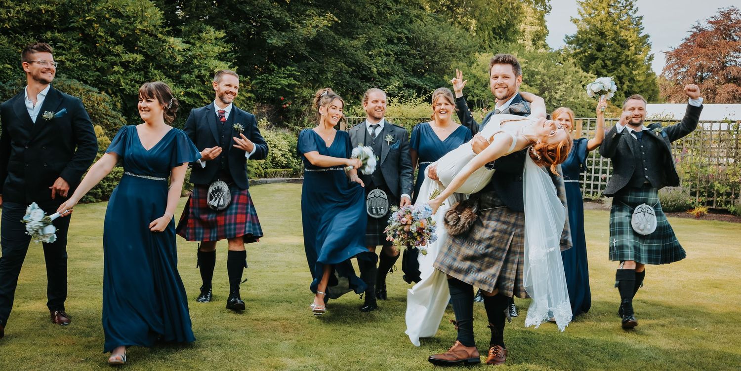 Wedding guests in matching blue dresses and traditional Scottish kilts dance joyfully on a sunny lawn.