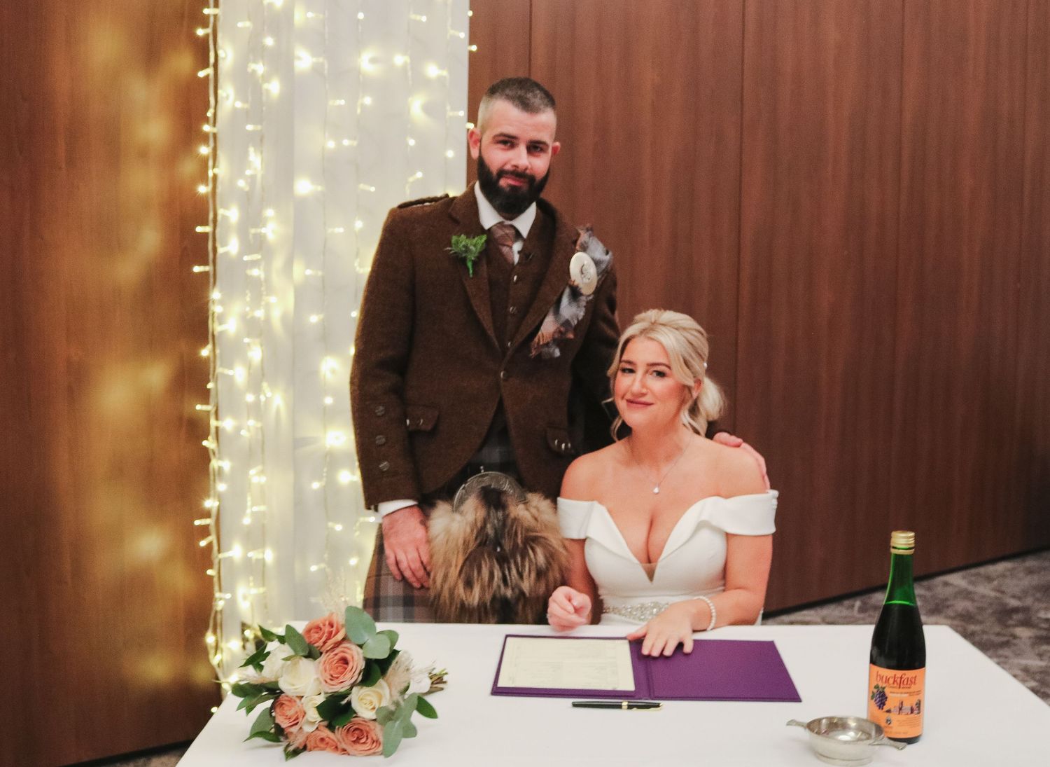A couple signs legal papers during a wedding ceremony with string lights and floral decor in the background.