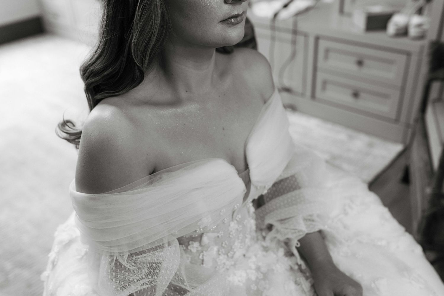 Black and white photo of person wearing delicate off-shoulder wedding dress with lace and tulle details.