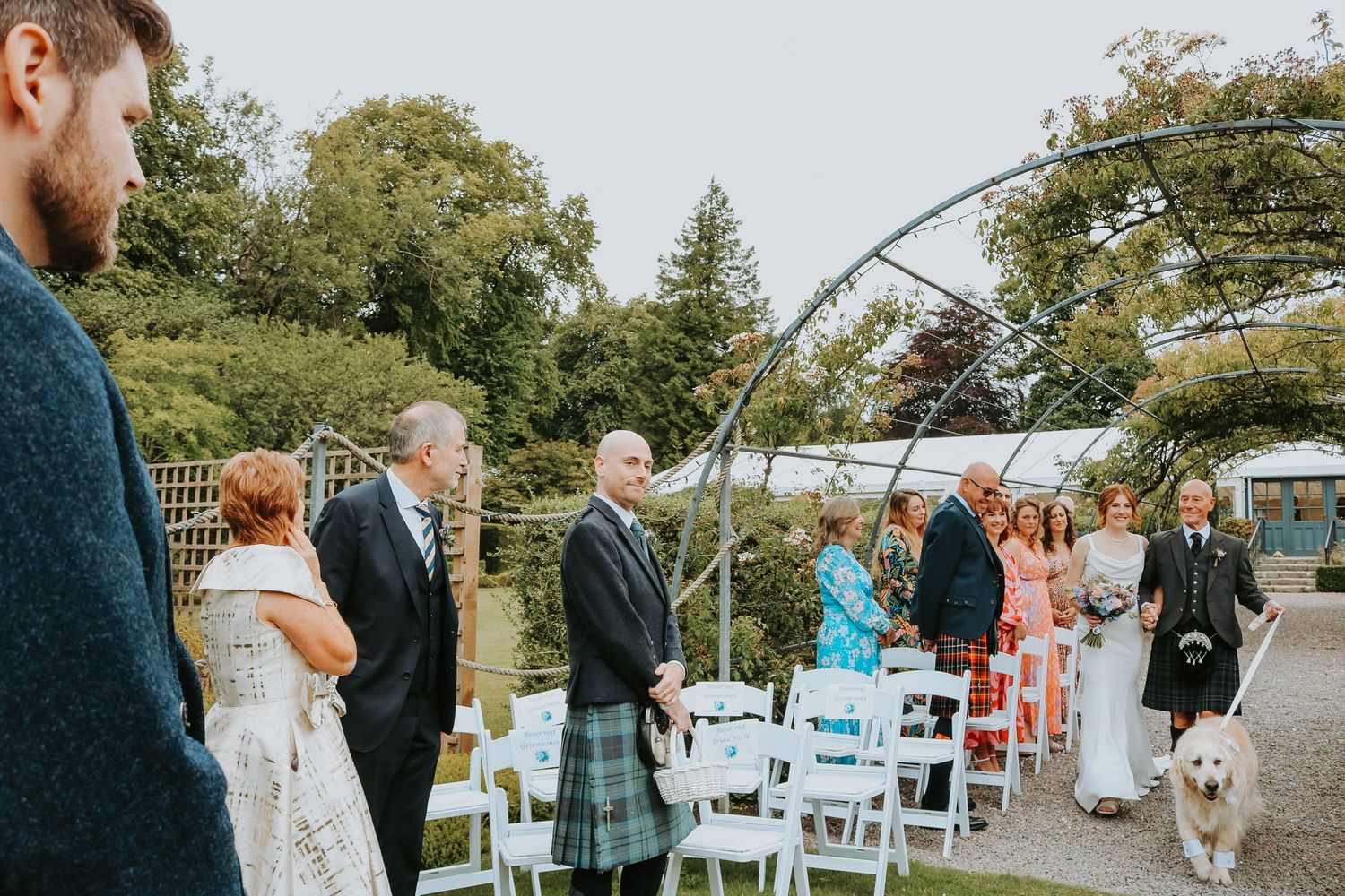 An outdoor wedding ceremony with guests seated on white chairs surrounded by trees and greenery on a sunny day.