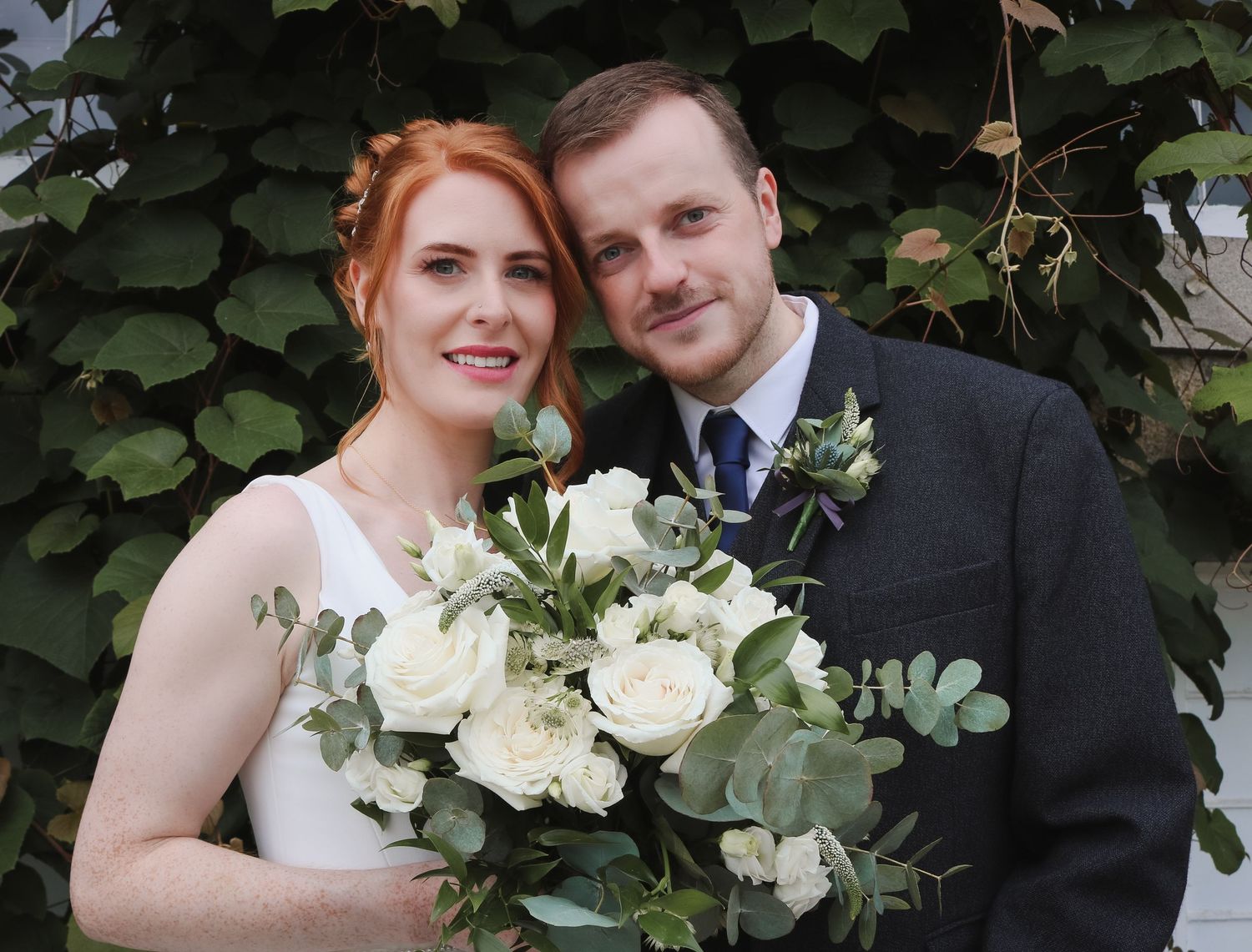 A romantic wedding portrait of a couple with a white floral bouquet against a leafy green backdrop.