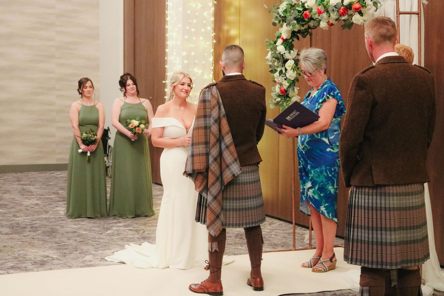 A wedding party stands in a formal line outside a church, with bridesmaids in green dresses and the groom in a kilt.