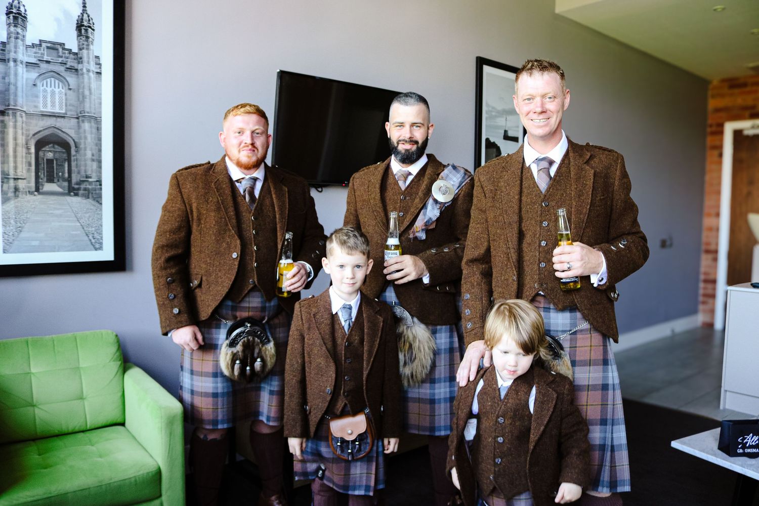 A group wearing coordinated brown tweed jackets and kilts poses together in a modern interior space with a green couch.