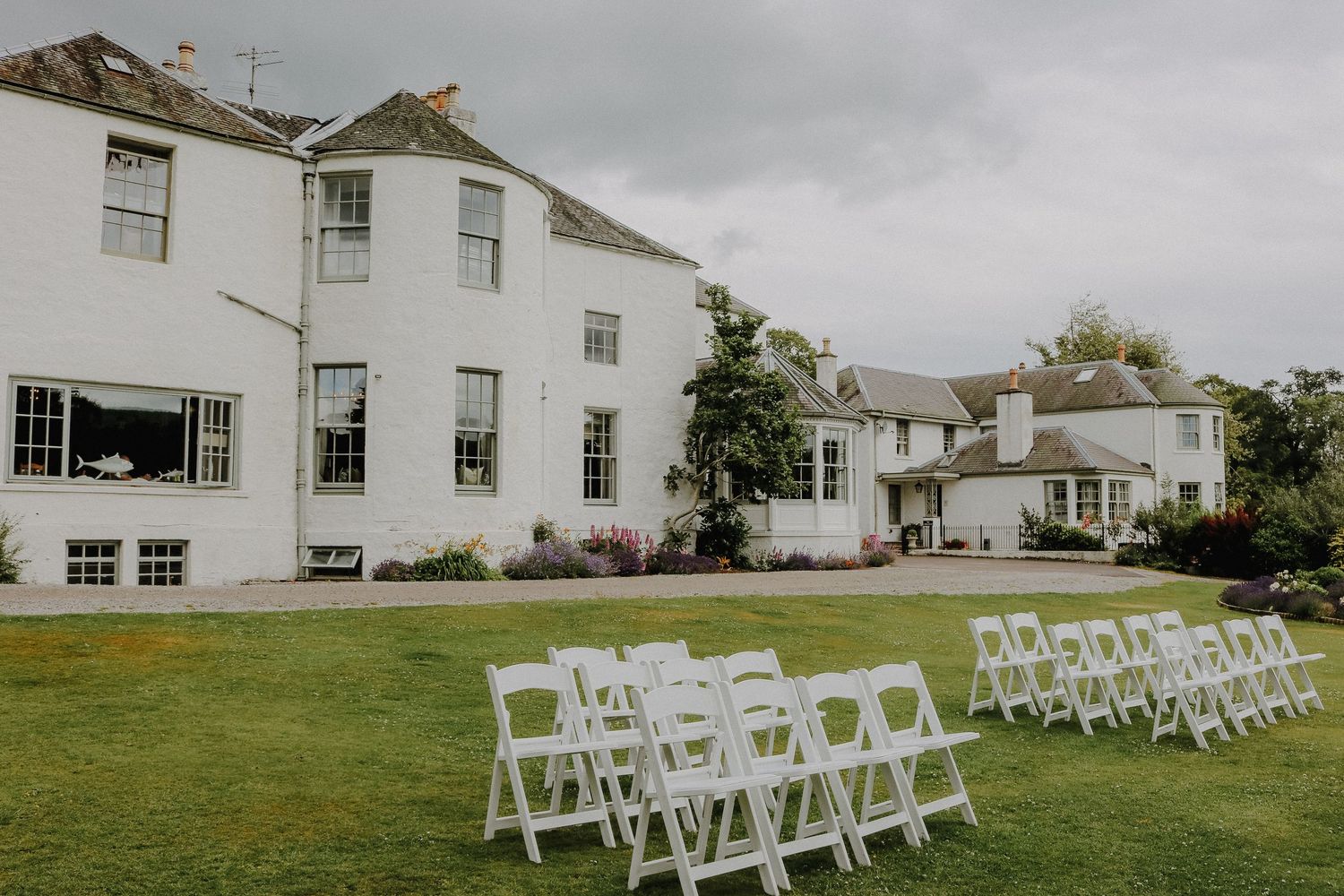 White folding chairs arranged in rows on a green lawn for an outdoor wedding ceremony with white buildings visible.
