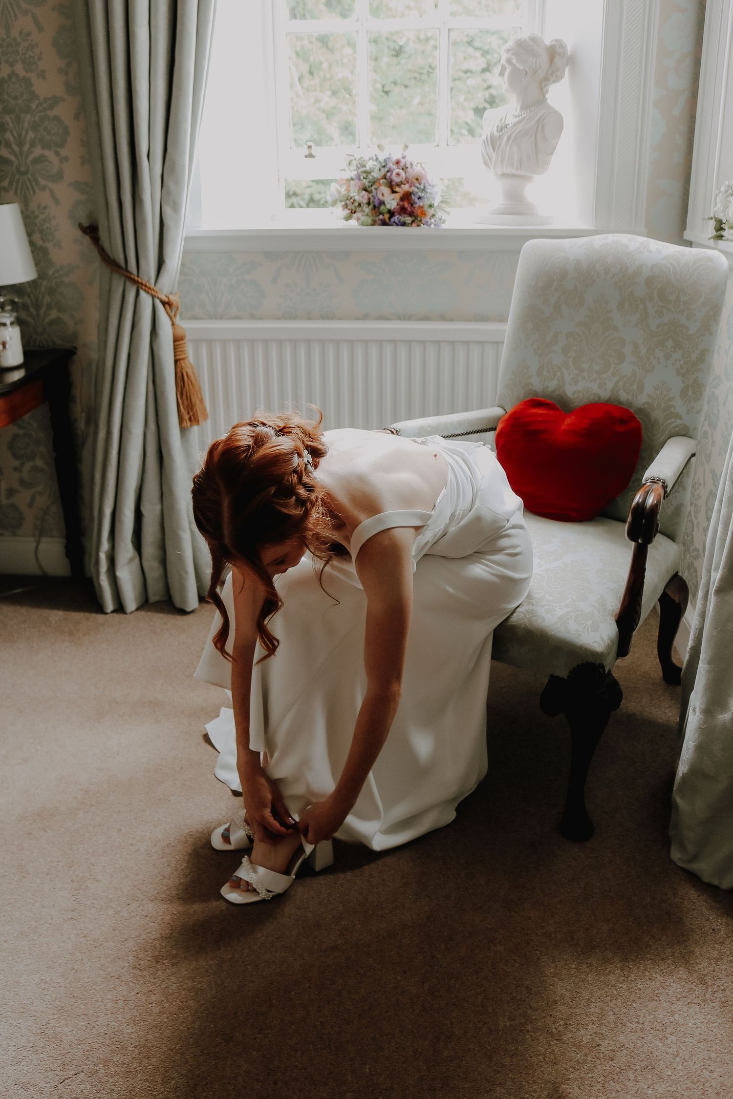 A person in a white dress fastens their shoes by a window with a red heart pillow on a nearby chair.