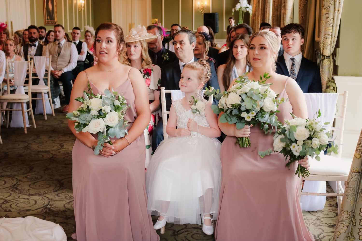 Several bridesmaids in soft pink dresses stand with bouquets waiting for the bride during an elegant wedding ceremony.