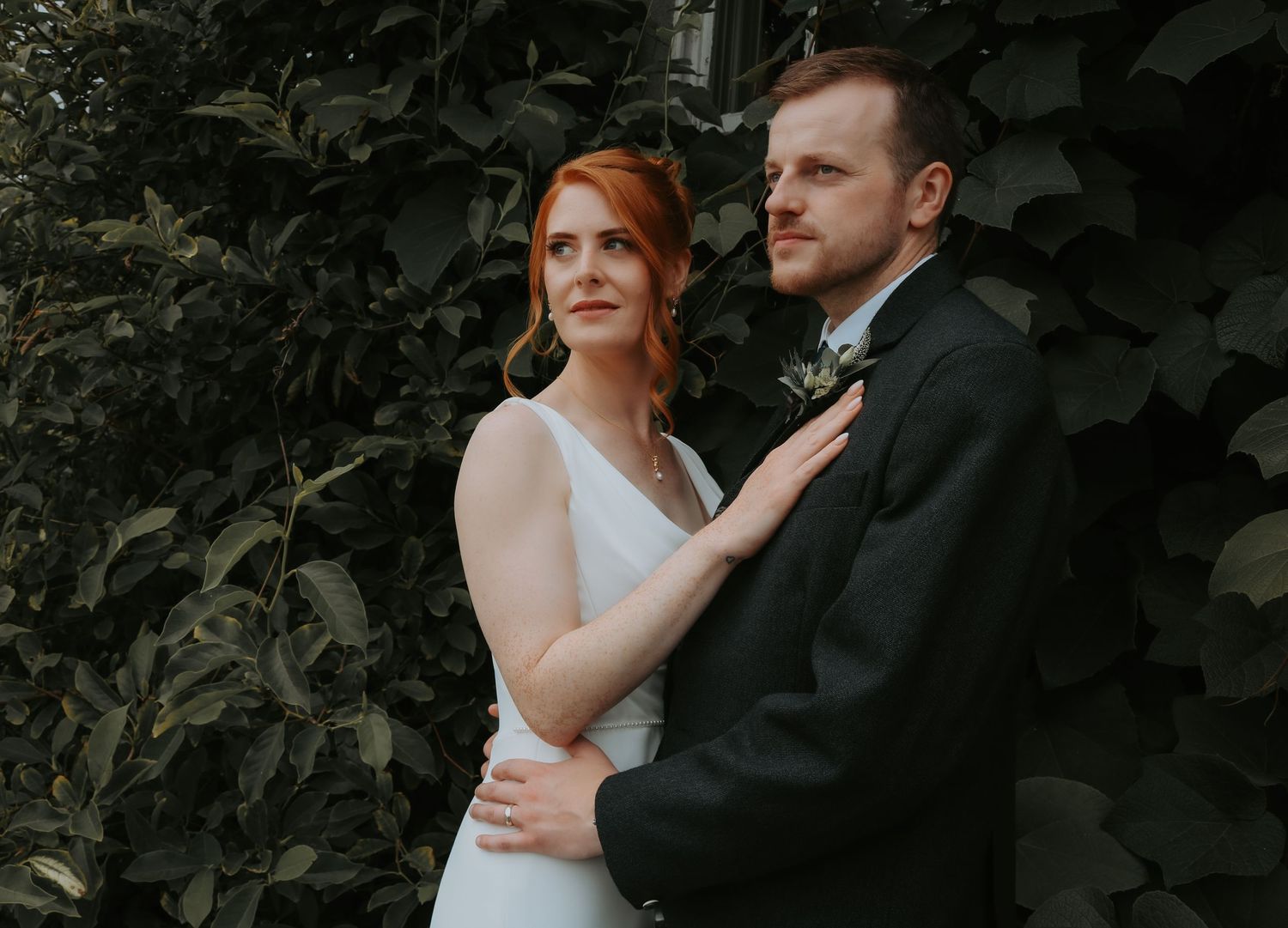 Newlywed couple embraces in front of lush green foliage during outdoor wedding portrait session.