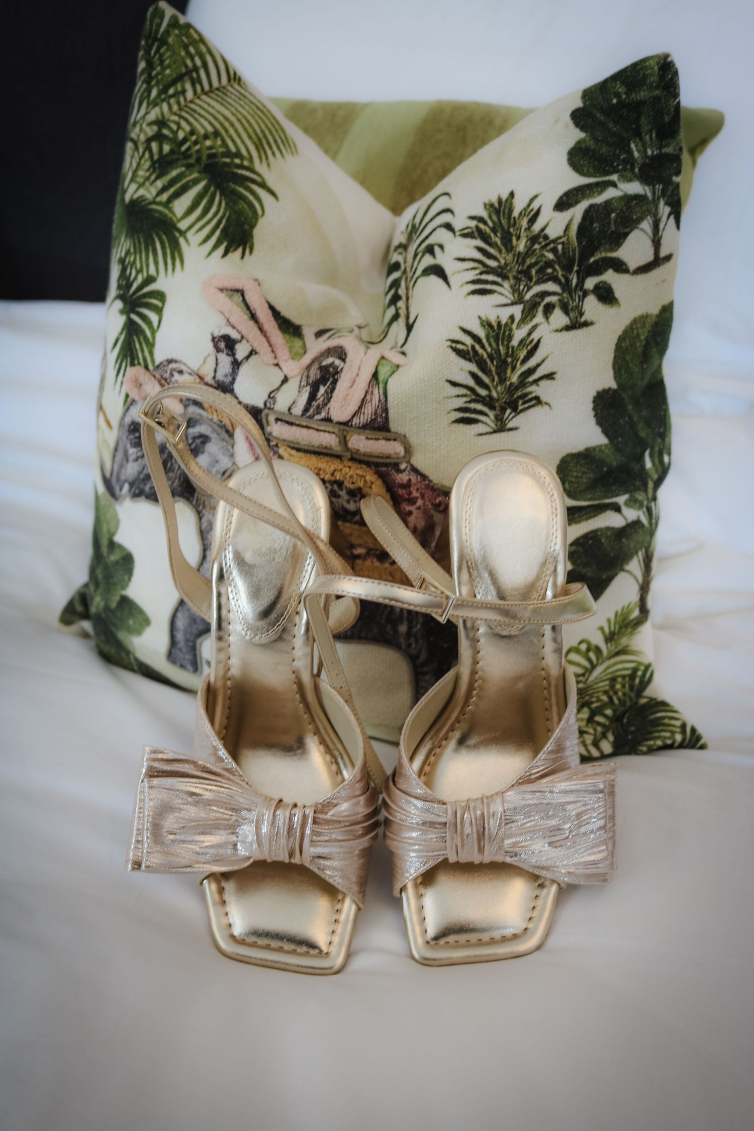 Metallic rose gold bow wedding sandals displayed at different angles against palm print fabric pattern cushions.
