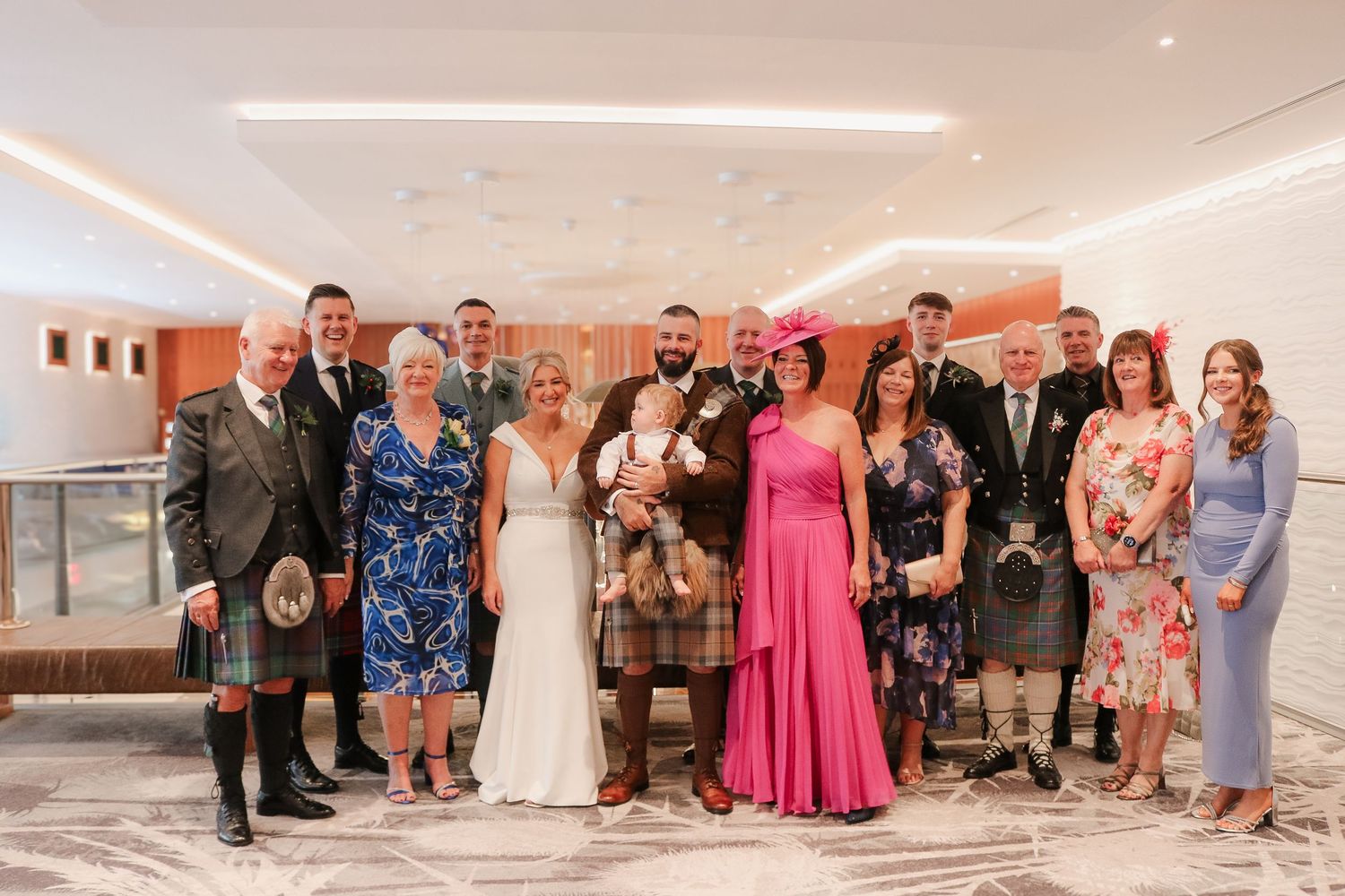 A wedding party group photo in formal attire standing together in an elegant indoor venue with soft lighting.