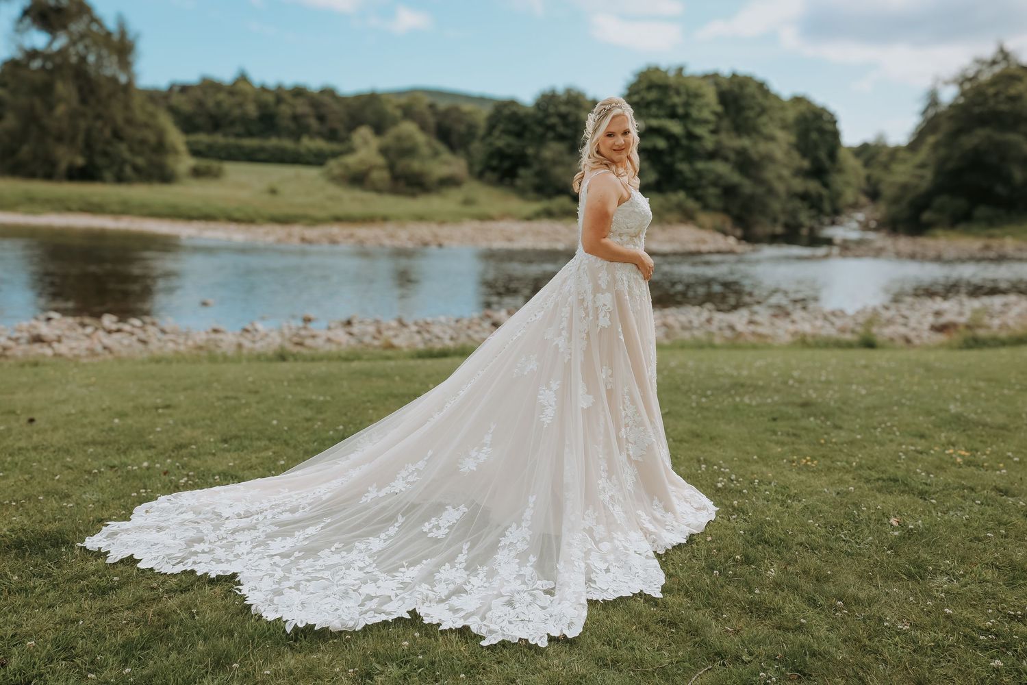 Elegant off-shoulder lace wedding dress with flowing train captured by lakeside on grassy lawn under blue sky.